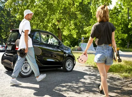 stadtmobil steht am Skateplatz, junge Erwachsene laufen mit Skateboard und Helm in der Hand Richtung Skateplatz