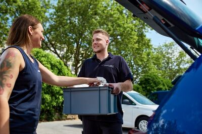 Mitarbeiter von stadtmobil hält eine Kiste mit Arbeitsutensilien in der Hand und Mitarbeiterin von stadtmobil greift in die Kiste um Arbeitsmaterial herauszuholen